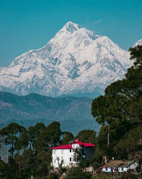 Snow-covered Himalayan peak rising above pine forest and a red-roofed hillside home near The MS Hills
